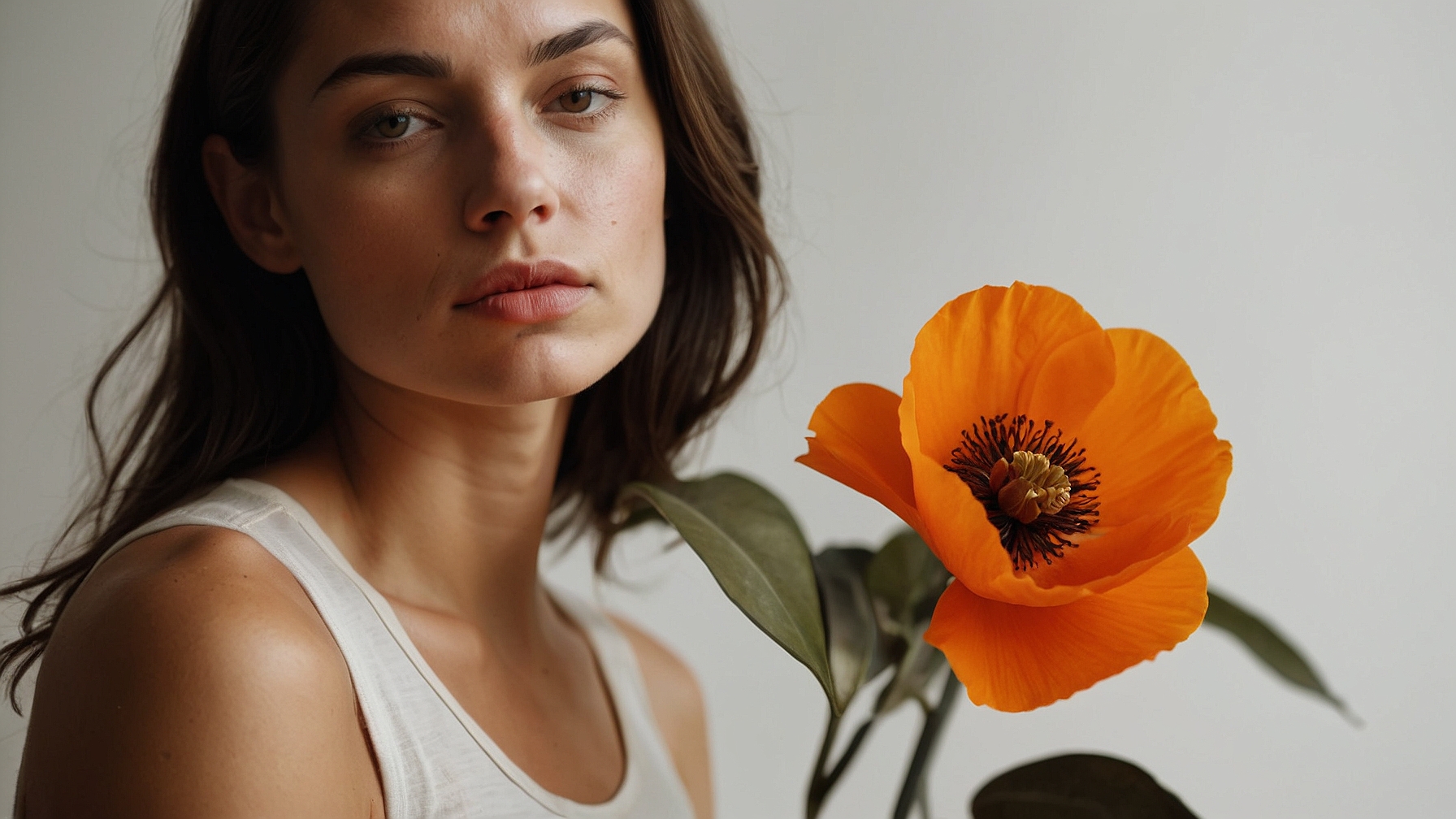Attractive woman with dark hair holding orange poppy flower in front of face, wearing white top, natural light, white background.