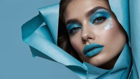 Portrait of woman with blue eyeshadow and turquoise lipstick posing in front of origami backdrop, wearing shades of sky blue.