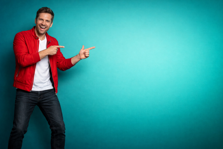 Smiling man in red jacket and white t-shirt enthusiastically pointing both hands to empty turquoise blue background space