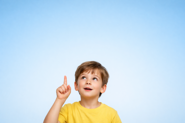 Young boy wearing yellow shirt pointing his index finger upward against a clear light blue background, looking intrigued