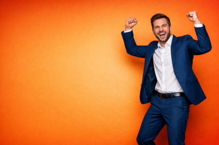 Happy young businessman in blue suit cheering with fists raised in front of an orange background