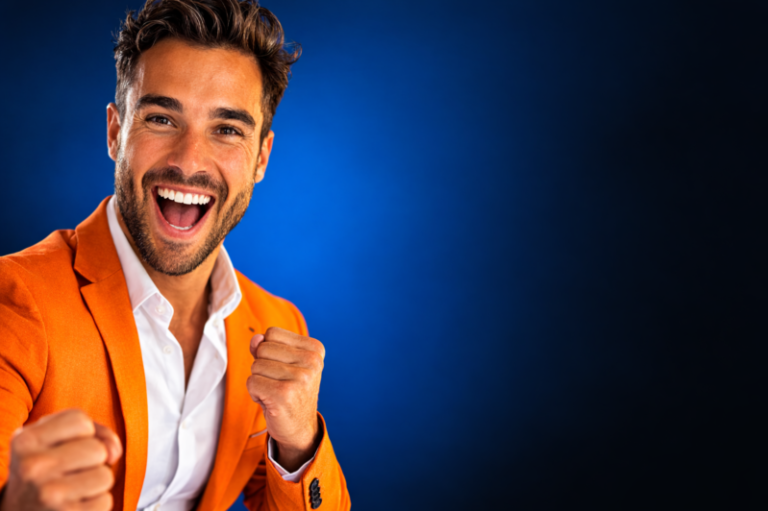 A young man with a joyful expression and clenched fists wearing a bright orange blazer against a blue background