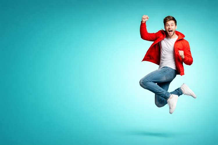 Joyful young man wearing a red jacket and blue jeans jumping in the air over a smooth blue background