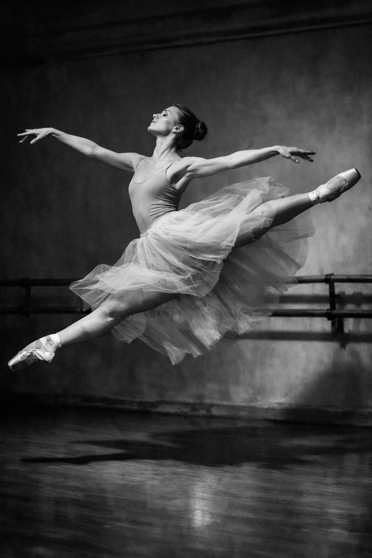 Black and white photo of a female ballet dancer performing a grand jeté with extended arms and legs in a studio