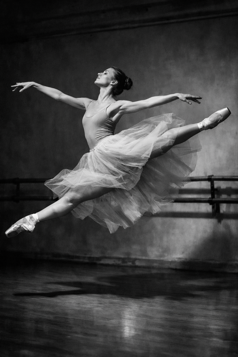 Black and white image of a female ballerina in mid-air executing a grand jeté in a dance studio