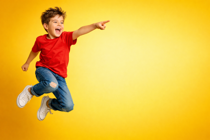 Happy young boy in red shirt and jeans energetically jumping and pointing with excitement against a bright yellow background