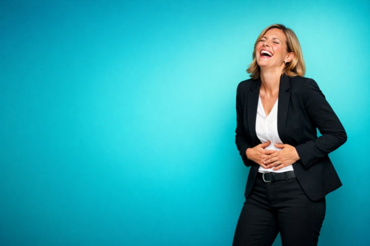 Smiling businesswoman wearing black suit laughing joyfully with hands on stomach against a blue studio background