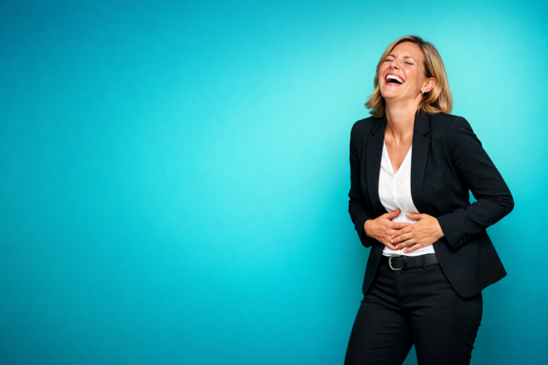 Smiling businesswoman wearing black suit laughing joyfully with hands on stomach against a blue studio background