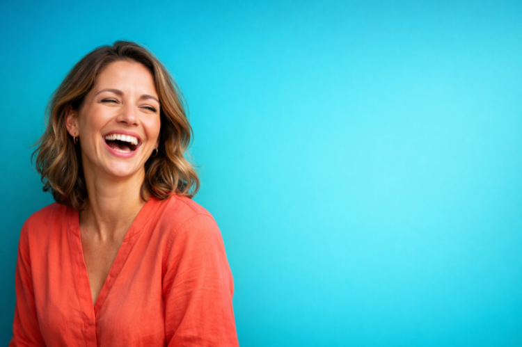 Portrait of a happy woman laughing joyfully wearing an orange shirt with light brown wavy hair on a blue background