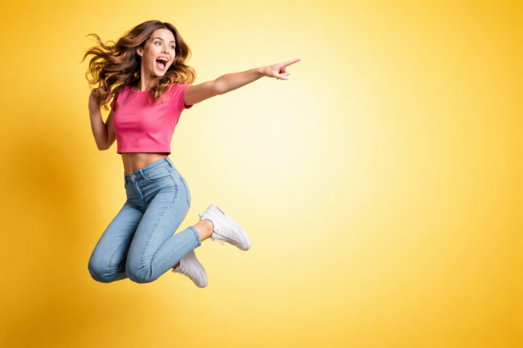 Happy young woman in a pink top and blue jeans jumping and pointing with excitement on a yellow background