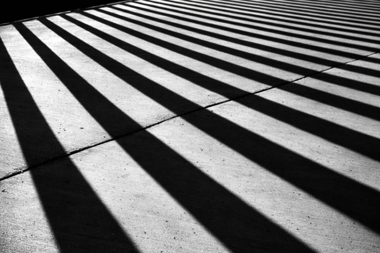 Black and white photo showing long, diagonal shadows of railings cast sharply on concrete pavement