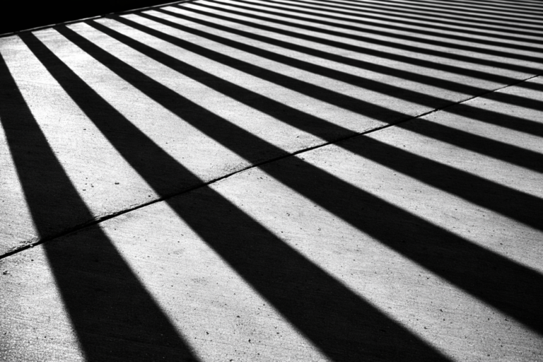 Black and white photo showing long, diagonal shadows of railings cast sharply on concrete pavement
