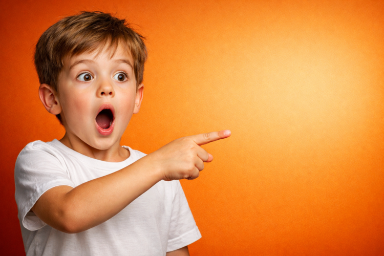 Young boy with light brown hair and white shirt looking surprised and pointing to the right against an orange backdrop
