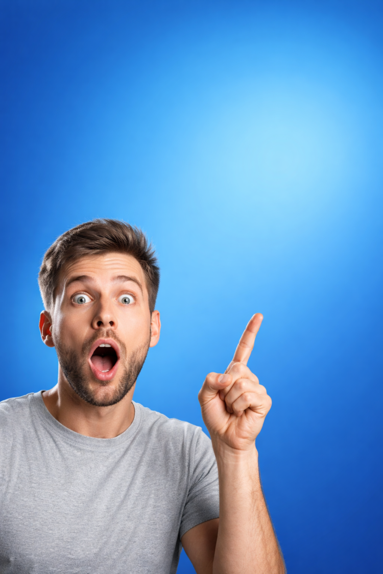 A young man with a beard and light gray t-shirt looking surprised, pointing upward with his index finger against a bright blue background
