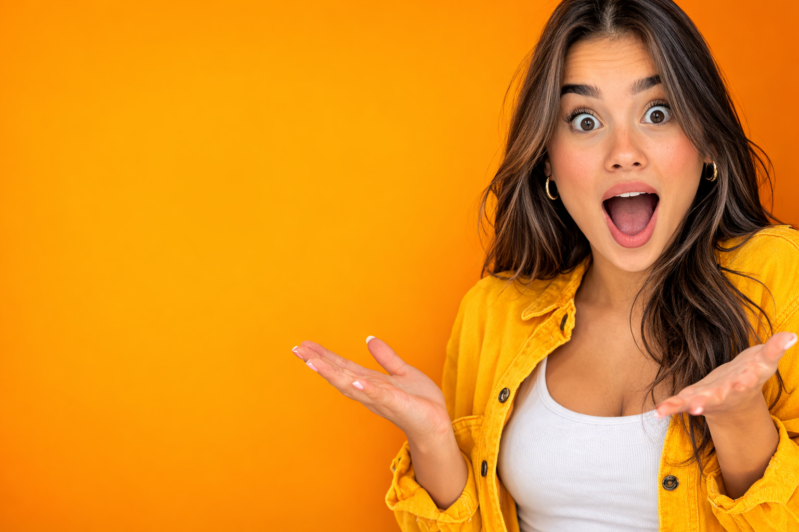 Young woman in mustard yellow jacket with wide eyes and open mouth showing surprise against solid bright orange background