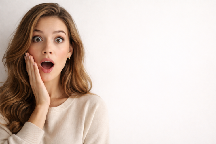 A shocked young woman with long wavy hair holds her cheek, eyes wide open and mouth agape, wearing a beige sweater against a plain background.
