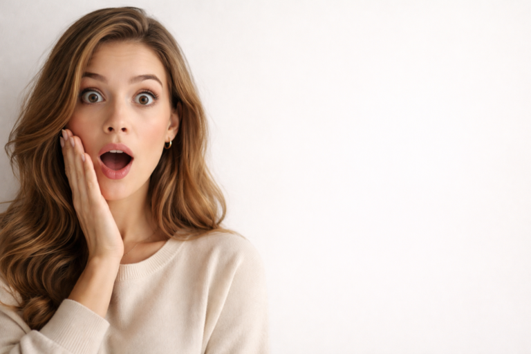 A shocked young woman with long wavy hair holds her cheek, eyes wide open and mouth agape, wearing a beige sweater against a plain background.