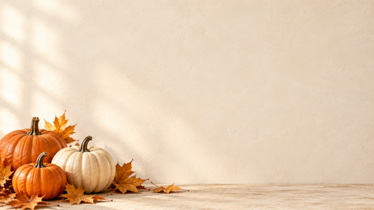 Several orange and white pumpkins arranged with autumn leaves on a wooden surface against a beige textured wall