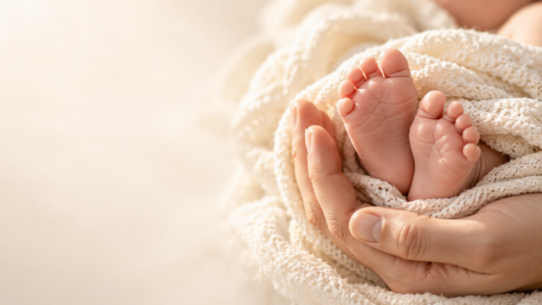 Close-up of tiny newborn baby feet held securely in adult hands wrapped in soft creamy knit fabric